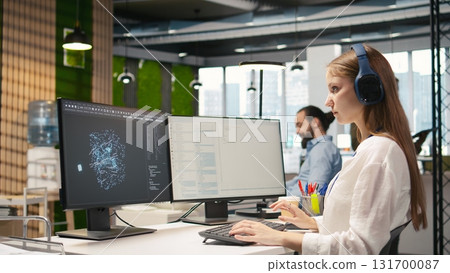 Female technician coding with AI language model assistance in startup office, listening music. Young woman wearing headphones integrating LLM assistant suggestions into code editor, camera A 131700087