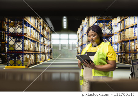 African american staff in workwear uses device to manage product handling, controlling inventory in warehouse with storage racks. Parcels organized for local delivery, maintaining high standards. 131700115