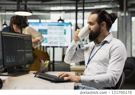 Employee sipping coffee while he reviews analytics and KPI metrics on monitor, driving business strategy and maintaining a professional mindset focused on objectives and success. 131700178