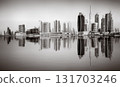 Monochrome city skyline with tall skyscrapers reflecting on calm water under a cloudy sky, Dubai Marina bay UAE 131703246