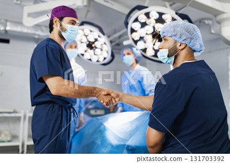 Two surgeons shake hands in an operating room, with other medical staff and surgical equipment in the background. Two surgeons shake hands in an operating room, with other medical staff and surgical equipment in the background. 131703392