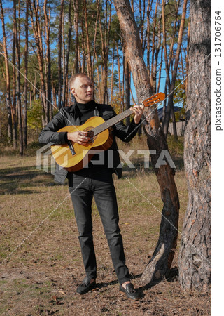 Portrait of smiling caucasian artist plays wooden acoustic guitar in autumnal park. Young songwriter plays string musical instrument hobby outside in nature fall time. Audio music healing Portrait of smiling caucasian artist plays wooden acoustic guitar in autumnal park. Young songwriter plays string musical instrument hobby outside in nature fall time. Audio music healing 131706764