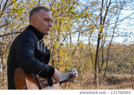Beautiful blonde man playing acoustic guitar and singing relaxing enjoying outdoors on sunny autumn day. Artist learning and practicing playing musical instrument. Fall leaves autumnal Beautiful blonde man playing acoustic guitar and singing relaxing enjoying outdoors on sunny autumn day. Artist learning and practicing playing musical instrument. Fall leaves autumnal 131706803