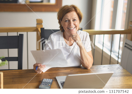 happy senior woman calculating family budget, sitting at kitchen counter with laptop, papers, 131707542