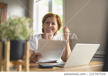 happy senior woman calculating family budget, sitting at kitchen counter with laptop, papers, 131707544