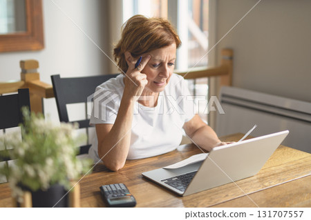 Sad frustrated senior woman pensioner having depressed look, holding hand on her face, calculating family budget, sitting at kitchen counter with laptop, papers, coffee, calculator and cell phone 131707557