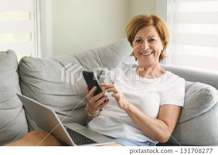 senior woman relaxing at home on the sofa reading her text messages on her mobile phone with a quiet smile 131708277
