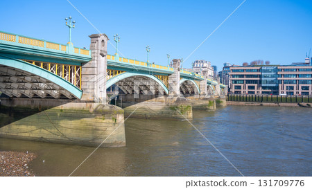 Southwalk Bridge stretches over the River Thames, showcasing its impressive structure. The clear blue sky reflects on the water, highlighting the bridge's beauty and nearby buildings. Southwalk Bridge stretches over the River Thames, showcasing its impressive structure. The clear blue sky reflects on the water, highlighting the bridge's beauty and nearby buildings. 131709776