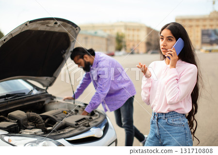 Frustrated Indian woman calling for help while man checks broken car engine 131710808