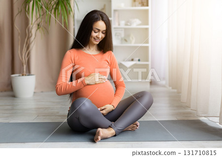 Pregnant woman sitting on yoga mat at home and touching belly 131710815