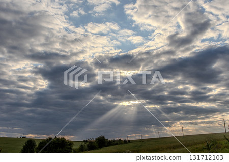 Rays of sunlight breaking through clouds over a rural landscape at sunset casting a serene glow across the fields and hills Rays of sunlight breaking through clouds over a rural landscape at sunset casting a serene glow across the fields and hills 131712103