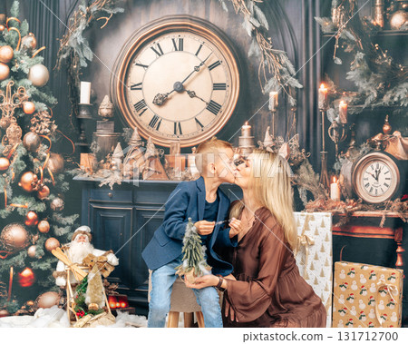 Tender moment of mother kissing her son near Christmas fireplace. Cozy home atmosphere with decorated tree, lights, and presents 131712700
