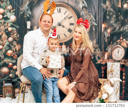 Festive family portrait with parents and child sitting near tree and presents. Smiling faces and cozy studio lights capture warmth and joy 131712701