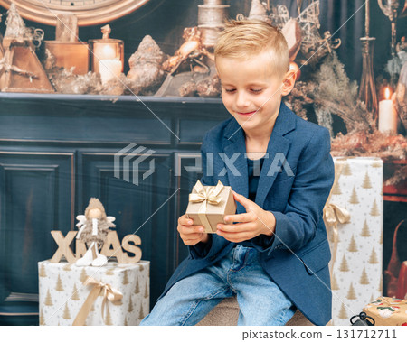 Smiling boy opening Christmas gift near holiday decorations, lights and wrapped boxes. Joyful festive emotion Smiling boy opening Christmas gift near holiday decorations, lights and wrapped boxes. Joyful festive emotion 131712711