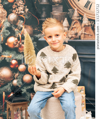Little boy smiles while holding small decorative tree in festive studio with warm lights and cozy atmosphere Little boy smiles while holding small decorative tree in festive studio with warm lights and cozy atmosphere 131712712