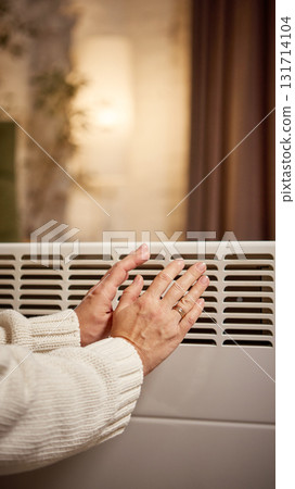 Hands warming near electric convector in softly lit living room 131714104