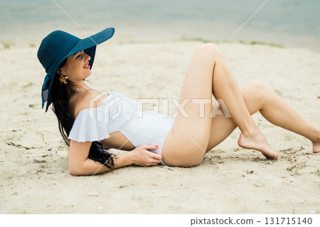 A girl in a white swimsuit is lying on the warm sand, adorned with a blue hat A girl in a white swimsuit is lying on the warm sand, adorned with a blue hat 131715140