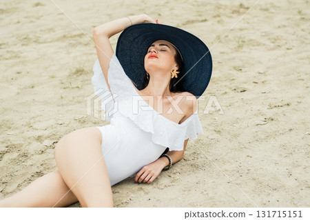 A young woman in a white swimsuit is sitting on the beach with a blue hat 131715151