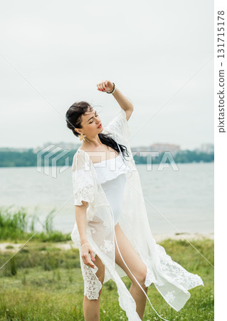 A girl is standing on the sand near a lake in a white swimsuit and a light pareo A girl is standing on the sand near a lake in a white swimsuit and a light pareo 131715178