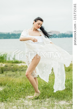 A young woman poses on the shore near clear water in an elegant white outfit 131715181