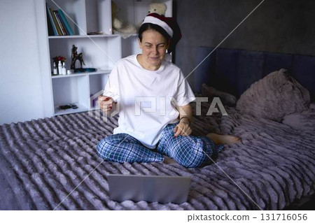 a senior woman in a Santa Claus hat and with a sparkler on a video conference wishes everyone a Merry Christmas at home via the Internet 131716056