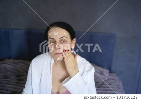 a senior woman in underwear on the bed performs routine self-care with face and body cosmetics 131716122