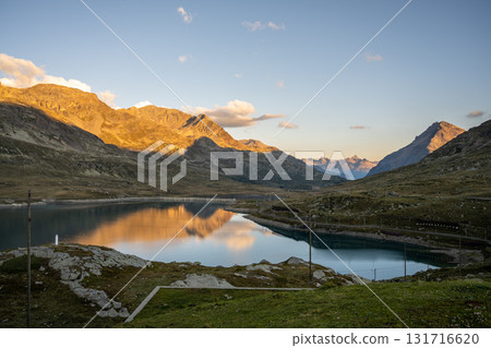 Lago Bianco glimmers under the rising sun at Bernina Pass in Graubunden. The surrounding mountains reflect on the tranquil waters, showcasing the natural beauty of the Swiss Alps. 131716620