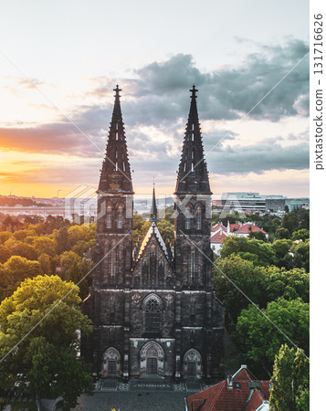 The majestic Vysehrad Basilica of St. Peter and St. Paul stands tall amidst lush greenery as the sun rises over Prague, casting a warm glow on its impressive architecture and surroundings. 131716626