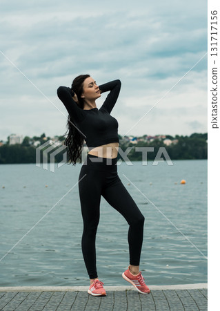 A girl with an athletic build in dark sportswear poses against the backdrop of a lake 131717156