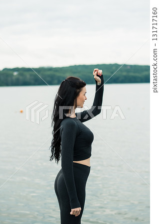 A young woman with an athletic build is sitting on the shore by the water A young woman with an athletic build is sitting on the shore by the water 131717160