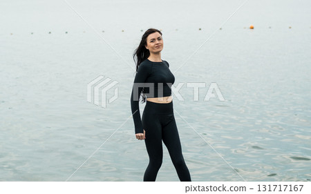 A young woman with an athletic build and long black hair poses near the lake shore 131717167