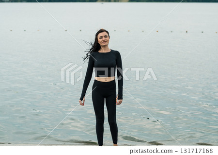 A young woman with an athletic build and long black hair poses near the lake shore A young woman with an athletic build and long black hair poses near the lake shore 131717168
