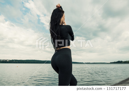A young girl with an athletic build stands against the backdrop of a lake in black leggings and a sweatshirt 131717177