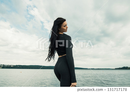 A young girl with an athletic build stands against the backdrop of a lake in black leggings and a sweatshirt 131717178