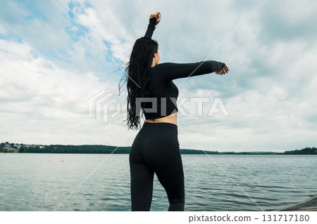 An athlete in dark sportswear poses on the shore of a lake surrounded by nature 131717180