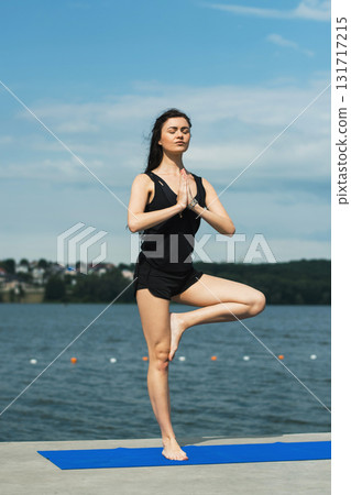 A young girl with an athletic build performs a yoga asana on the shore of a lake A young girl with an athletic build performs a yoga asana on the shore of a lake 131717215