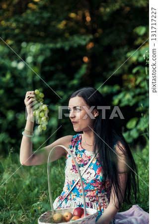 A girl at a picnic in the park holds a bunch of grapes against a backdrop of green trees 131717227