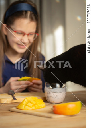 Black cat drinks chia pudding from a glass while girl smiles holding a piece of mango 131717688