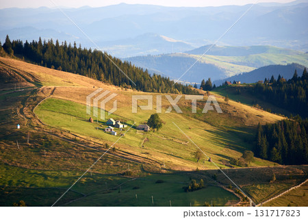 Expansive rural landscape in late afternoon light, featuring gently sloping hills and small farmhouse nestled in green meadow. Scene framed by dense forests and distant mountain ranges. 131717823