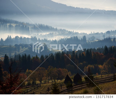 Morning sunlight streams through mist, illuminating serene mountain village nestled among forested hills. Light creates ethereal rays that gently touch autumn foliage and houses scattered on slopes. 131717834
