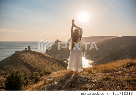 woman stands on a hill overlooking the ocean, her arms raised in the air. Concept of freedom and joy, as if the woman is celebrating a moment of happiness or accomplishment. 131718318