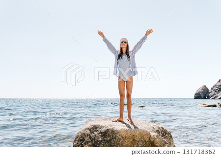 Woman Beach Joy: Lady raising arms on rock by sunny sea for summer freedom, vacation happiness. 131718362
