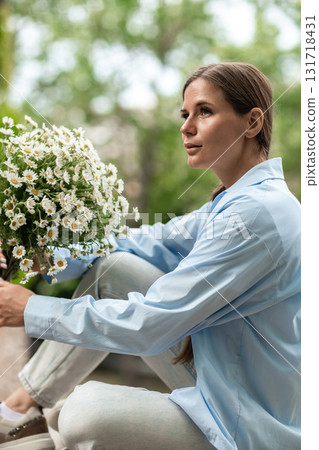 Woman Daisies Relaxation: Contemplative lady with daisies in park, sits on stone, spring day, enjoying nature's beauty. 131718431