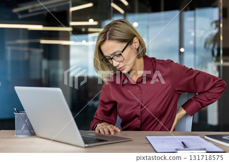 Businesswoman with eyeglasses experiencing back pain while working on laptop in a modern office, touching her back with hand and showing painful expression Businesswoman with eyeglasses experiencing back pain while working on laptop in a modern office, touching her back with hand and showing painful expression 131718575