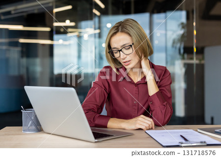 Stressed businesswoman is massaging her neck while working on a laptop in a modern office, experiencing discomfort and tension due to prolonged computer use 131718576