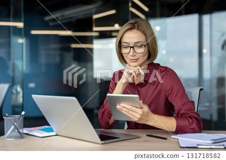 Smiling businesswoman engaging with a digital tablet while working on a laptop at her modern office desk, enjoying a productive and efficient workday filled with creativity and focus Smiling businesswoman engaging with a digital tablet while working on a laptop at her modern office desk, enjoying a productive and efficient workday filled with creativity and focus 131718582