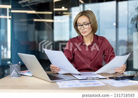 Focused businesswoman wearing glasses reads documents at a modern office desk, surrounded by a laptop, charts, and financial reports, exuding professionalism and concentration Focused businesswoman wearing glasses reads documents at a modern office desk, surrounded by a laptop, charts, and financial reports, exuding professionalism and concentration 131718585
