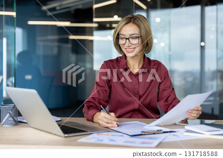 Happy businesswoman wearing glasses, reviewing documents and taking notes while working at a sleek desk in a modern office, surrounded by a laptop and organized paperwork Happy businesswoman wearing glasses, reviewing documents and taking notes while working at a sleek desk in a modern office, surrounded by a laptop and organized paperwork 131718588