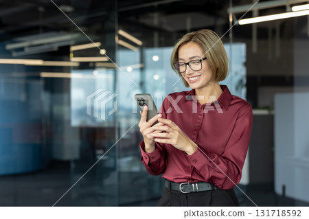 Happy businesswoman wearing glasses and elegant clothes is using smartphone in modern office, enjoying online communication and mobile technology for business Happy businesswoman wearing glasses and elegant clothes is using smartphone in modern office, enjoying online communication and mobile technology for business 131718592