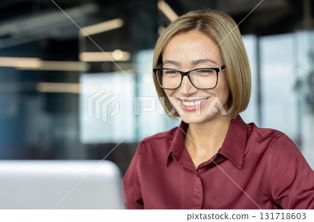 Happy businesswoman wearing eyeglasses, smiling while working on her laptop in a modern office, enjoying the rewards of her successful online business and feeling accomplished Happy businesswoman wearing eyeglasses, smiling while working on her laptop in a modern office, enjoying the rewards of her successful online business and feeling accomplished 131718603